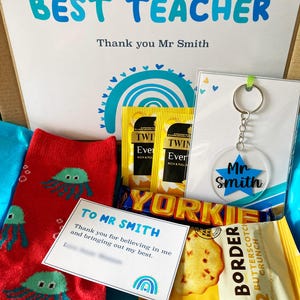 Könnte beinhalten: Eine Geschenkbox mit roten Socken mit türkisfarbenen Quallen, zwei Twinings-Teebeuteln, einem Yorkie-Schokoriegel, einer Packung Border Butterscotch Crunch-Keksen, einem Schlüsselanhänger mit blauem Stern Mr Smith und Karten mit Aufschriften.