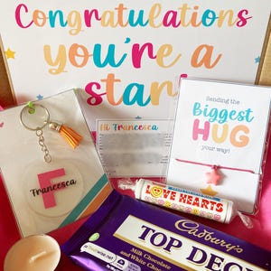 May include: A gift box filled with various items, including a chocolate bar, a keychain, a candle, and a card that says "Congratulations you're a star". The card is decorated with colourful stars. The chocolate bar is a Cadbury's Top Deck, a milk and white chocolate bar. The keychain is a clear acrylic circle with the letter "F" and the name "Francesca" written on it. The candle is a small, white tea light. The card also includes the phrase "Sending the Biggest Hug your way!"