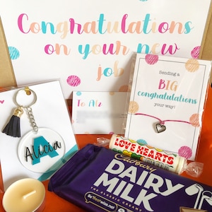 K&ouml;nnte beinhalten: Eine Geschenkbox gef&uuml;llt mit Artikeln zur Feier eines neuen Jobs. Die Box enth&auml;lt eine Schokoladentafel, einen Schl&uuml;sselanh&auml;nger mit einem personalisierten Namensschild, eine kleine Kerze, ein Armband und zwei Karten mit Gl&uuml;ckwunschbotschaften.