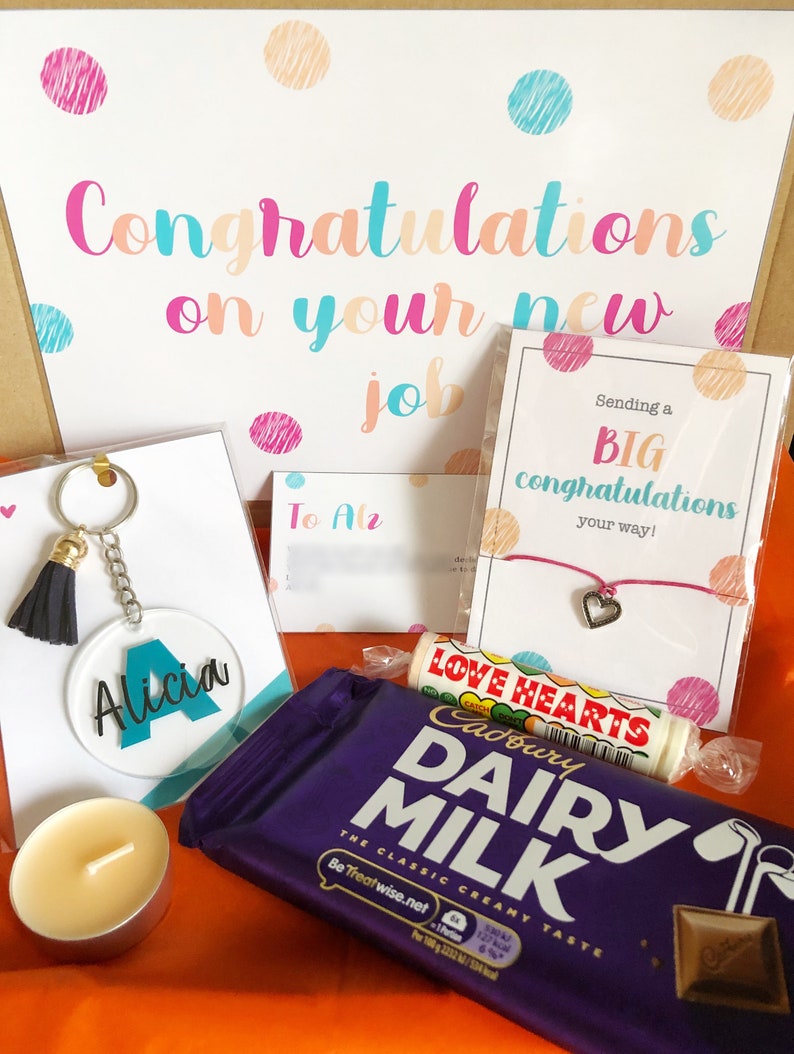 K&ouml;nnte beinhalten: Eine Geschenkbox gef&uuml;llt mit Artikeln zur Feier eines neuen Jobs. Die Box enth&auml;lt eine Schokoladentafel, einen Schl&uuml;sselanh&auml;nger mit einem personalisierten Namensschild, eine kleine Kerze, ein Armband und zwei Karten mit Gl&uuml;ckwunschbotschaften.