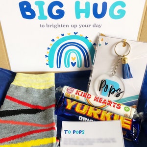 以下が含まれることがあります： 青い白のギフトボックスには、「Sending you a BIG HUG to brighten up your day」と書かれたカードが入っています。ボックスには、ストライプの靴下、チョコレートバー、キャンディーのチューブ、青いタッセルが付いたキーホルダーが入っています。