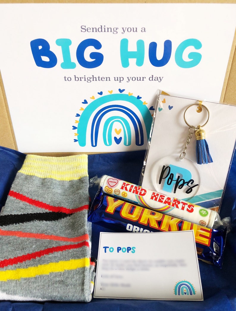 以下が含まれることがあります： 青い白のギフトボックスには、「Sending you a BIG HUG to brighten up your day」と書かれたカードが入っています。ボックスには、ストライプの靴下、チョコレートバー、キャンディーのチューブ、青いタッセルが付いたキーホルダーが入っています。
