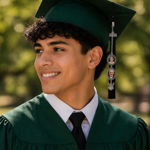 May include: A young person wearing a dark green graduation cap and gown. The cap has a tassel with a charm featuring the year 2026 and two small photo pendants. The person is smiling, wearing a white collared shirt and black tie.