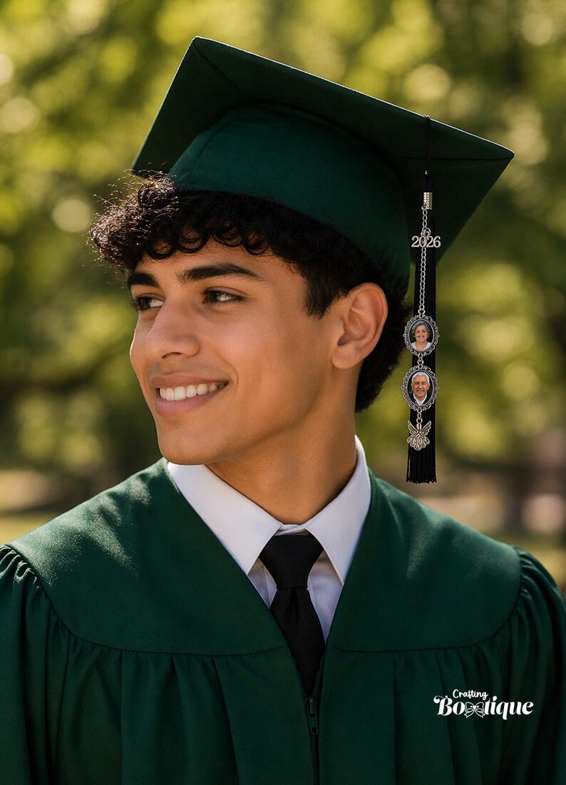 May include: A young person wearing a dark green graduation cap and gown. The cap has a tassel with a charm featuring the year 2026 and two small photo pendants. The person is smiling, wearing a white collared shirt and black tie.