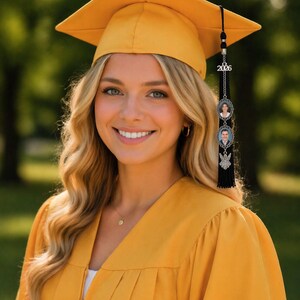 May include: A smiling person wearing a mustard yellow graduation cap and gown. The cap has a tassel with a charm that reads "2026" and includes two small photo pendants. The background is a blurred green outdoor scene.