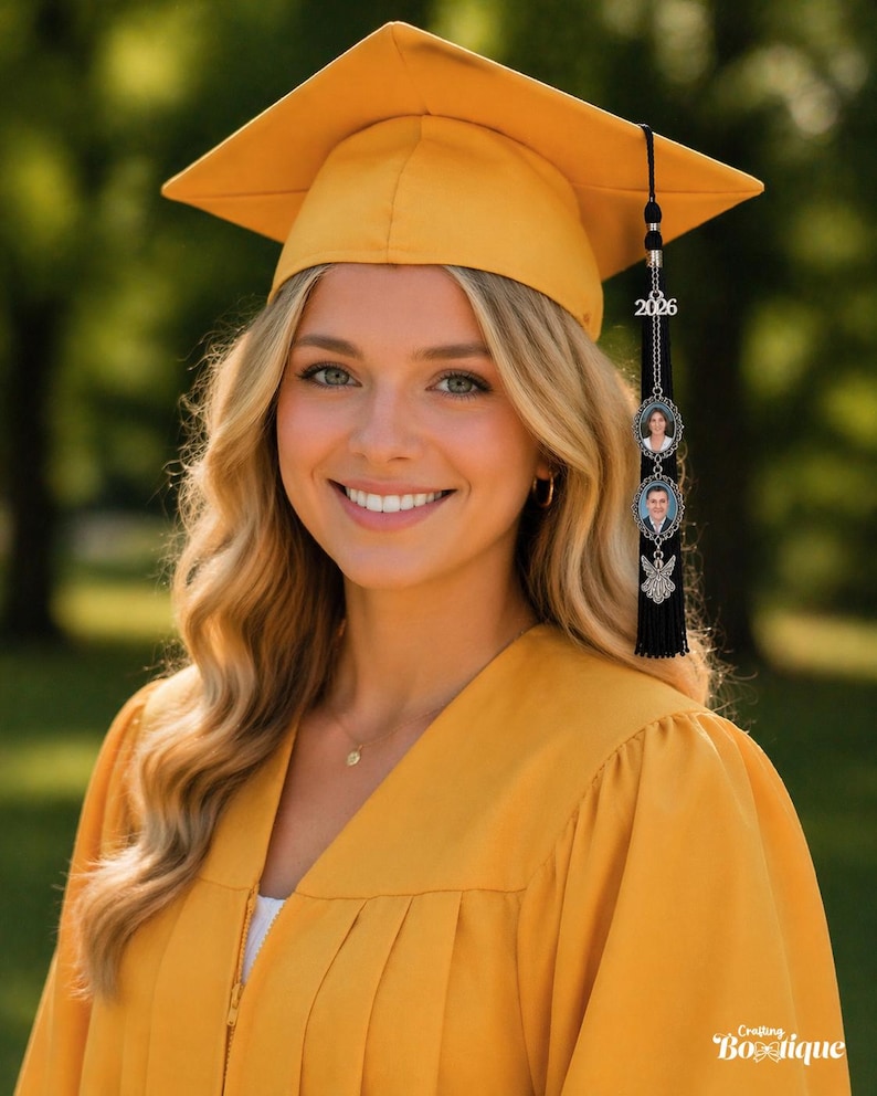 May include: A smiling person wearing a mustard yellow graduation cap and gown. The cap has a tassel with a charm that reads "2026" and includes two small photo pendants. The background is a blurred green outdoor scene.