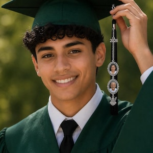 May include: A smiling graduate in a green cap and gown, holding a graduation tassel. The tassel features the year "2026" and two oval photo charms. The bottom of the tassel has a black fringe and a decorative charm. The image is set against a blurred green background.