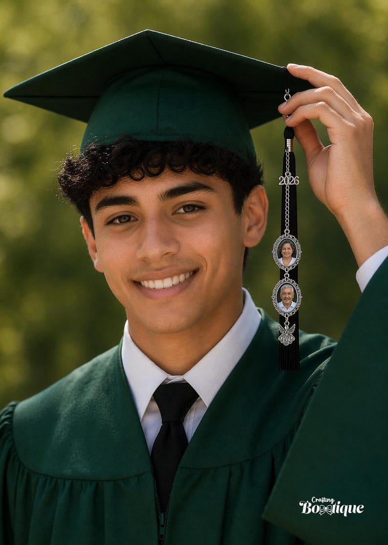 May include: A smiling graduate in a green cap and gown, holding a graduation tassel. The tassel features the year "2026" and two oval photo charms. The bottom of the tassel has a black fringe and a decorative charm. The image is set against a blurred green background.
