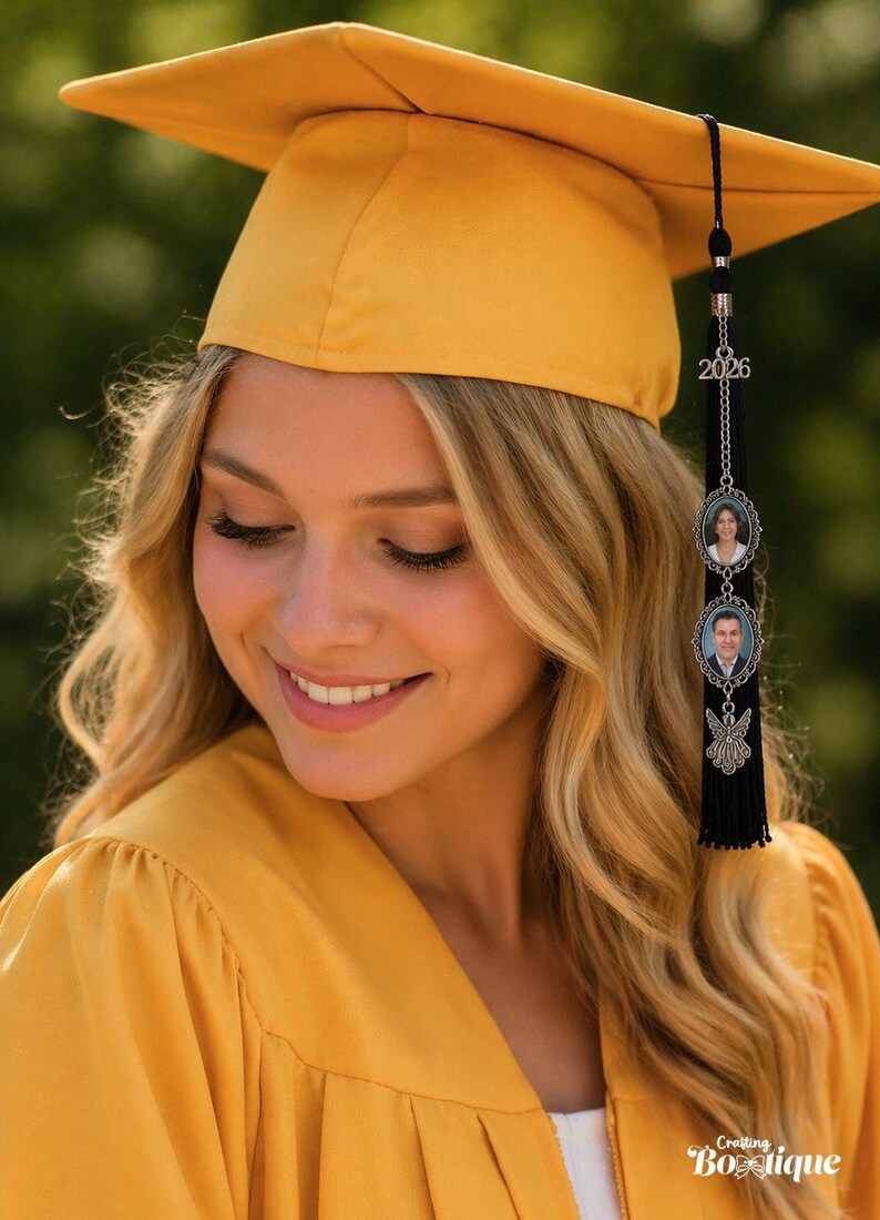 May include: A smiling person wearing a yellow graduation cap and gown. The cap has a black tassel with a charm featuring two oval portraits and the year 2026. The image is from Crafting Boutique.