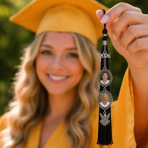 May include: A black graduation tassel with a silver charm featuring two oval photo frames and the year 2026. The tassel is held up in front of a person wearing a yellow graduation cap and gown.