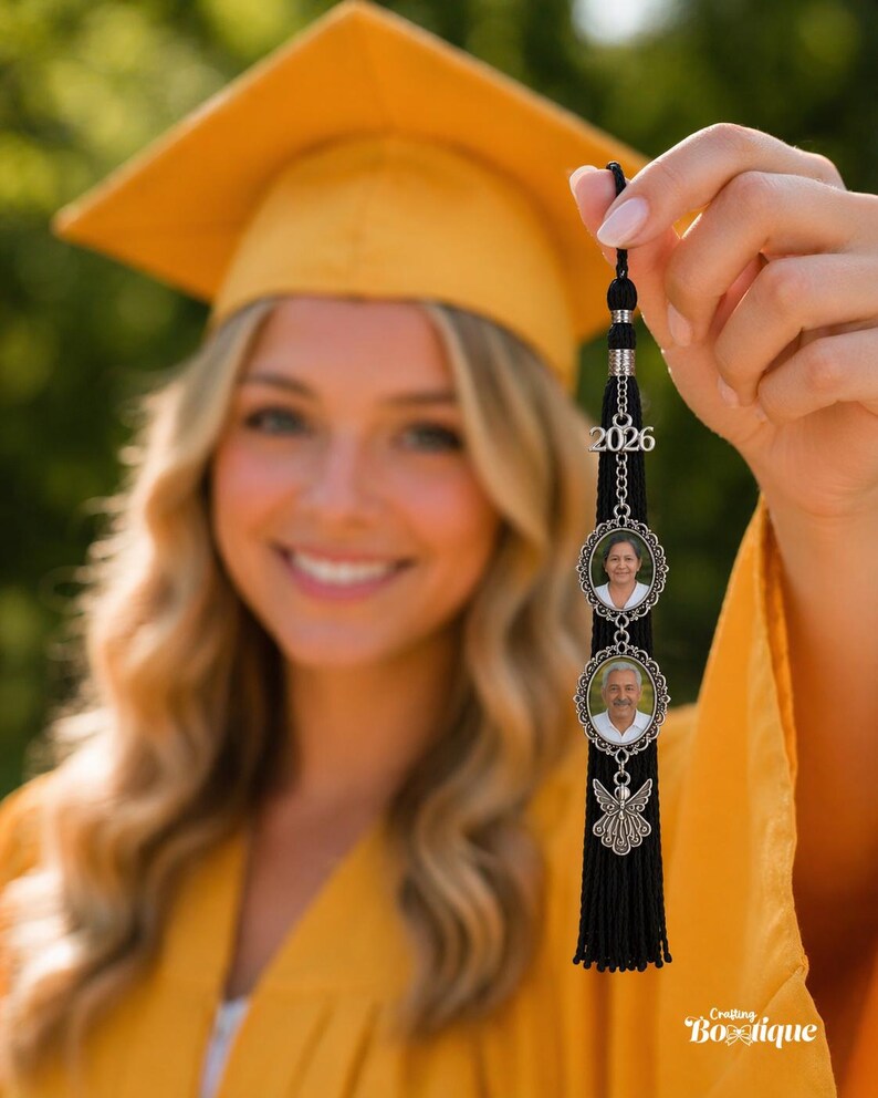 May include: A black graduation tassel with a silver charm featuring two oval photo frames and the year 2026. The tassel is held up in front of a person wearing a yellow graduation cap and gown.