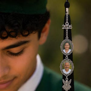May include: A black graduation tassel with a silver charm featuring the year "2026" and two oval photo frames. The frames hold small portraits. A decorative silver angel charm hangs below the photos. The image is of a person wearing a green graduation cap.