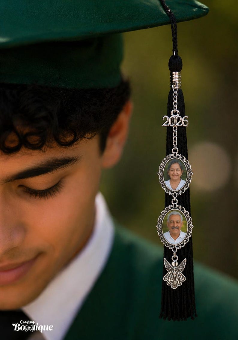 May include: A black graduation tassel with a silver charm featuring the year "2026" and two oval photo frames. The frames hold small portraits. A decorative silver angel charm hangs below the photos. The image is of a person wearing a green graduation cap.