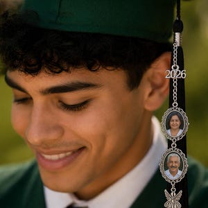 May include: A graduation tassel with a silver charm featuring the year 2026 and two oval photo frames. The tassel is black and hangs from a green graduation cap. The photo frames contain images of two people.