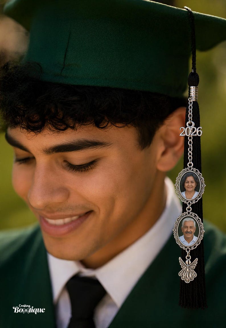 May include: A graduation tassel with a silver charm featuring the year 2026 and two oval photo frames. The tassel is black and hangs from a green graduation cap. The photo frames contain images of two people.