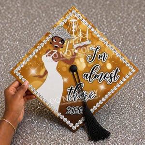 May include: A graduation cap with a gold and brown design, featuring a cartoon illustration of a woman in a white dress. The cap has the text "I'm almost there" and "2026". The edges are decorated with rhinestones and a black tassel.