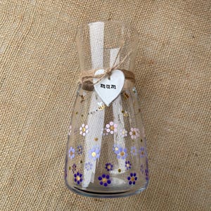 May include: A clear glass vase with a wide top and a narrow base. The vase is decorated with painted purple and pink flowers, gold dots, and bees. A heart-shaped tag with the word "mum" is tied around the neck with twine.