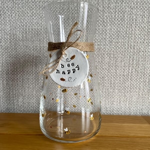 May include: A clear glass vase with a wide base and narrow neck, decorated with small gold and white bees. A twine band encircles the vase, holding a white circular tag that reads "bee happy" in black lettering.