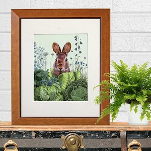 May include: A framed art print depicting a brown rabbit surrounded by green cabbages and blue flowers. The artwork is set against a pale green backdrop and framed in a wooden frame. A fern in a white pot is on the right.