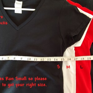 May include: A stack of five V-neck t-shirts in different sizes, from small to 3XL. The shirts are black, white, red, grey, and heather grey. A measuring tape is laid across the bottom of the shirts, with the corresponding sizes marked. The text "Sizes shown are small-3XL V Necks" is written in red at the top of the image. The text "Adult Ladies Sizes Run Small so please use measurement to get your right size." is written in red at the bottom of the image.