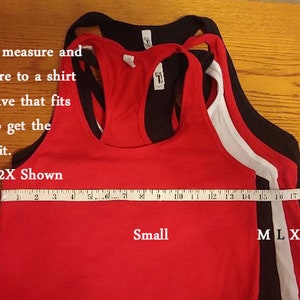 May include: Four tank tops in different sizes, laid out on a wooden surface. The tank tops are red, white, black, and black. A measuring tape is placed across the bottom of the tank tops, with the numbers 2 through 22 visible. The text "Small-2X Shown" is written above the measuring tape. The text "Small" is written below the red tank top. The text "M L XL 2XL" is written below the measuring tape.