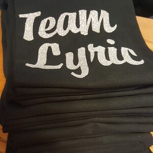 May include: A stack of black t-shirts with silver glitter lettering that reads "Team Lyric".