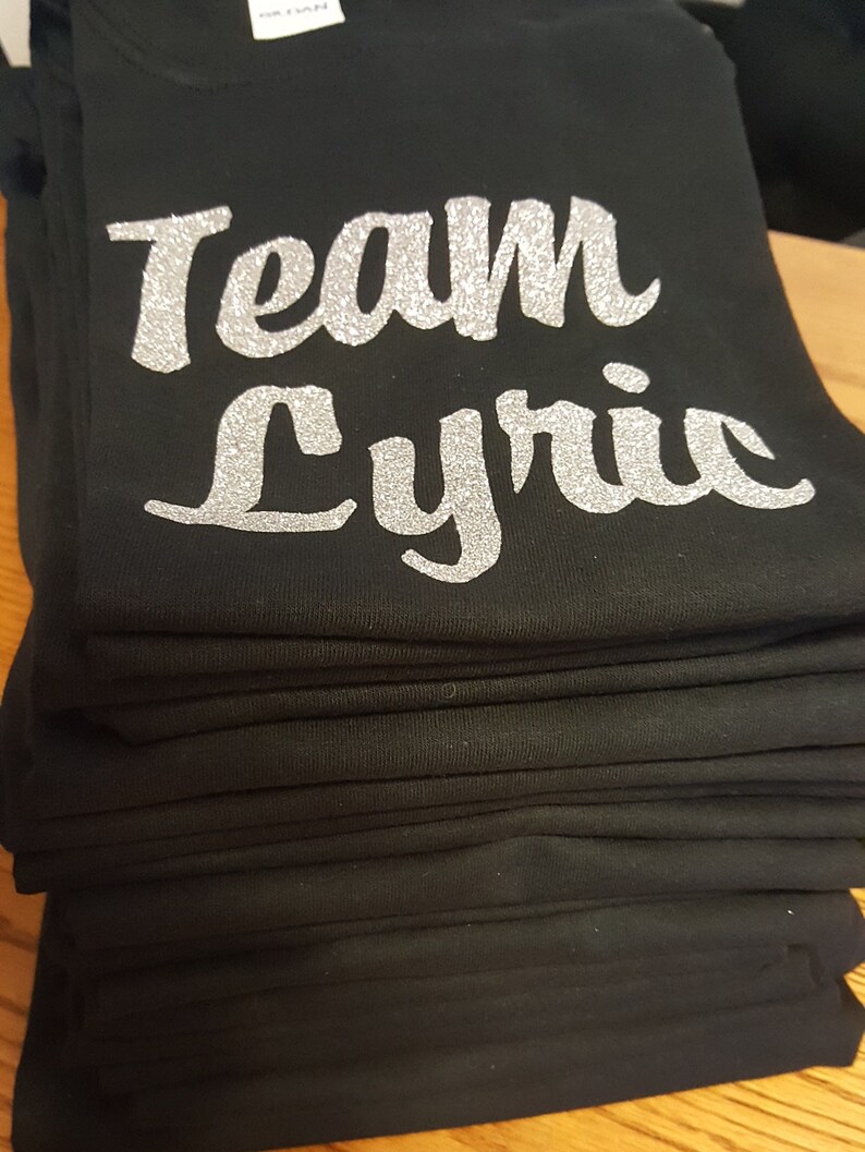 May include: A stack of black t-shirts with silver glitter lettering that reads "Team Lyric".