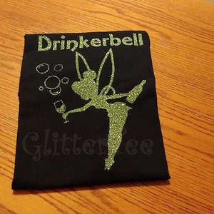 May include: Black t-shirt with a green glitter design of a fairy holding a glass of wine. The text "Drinkerbells" is above the fairy and "Glitterlee" is below the fairy.