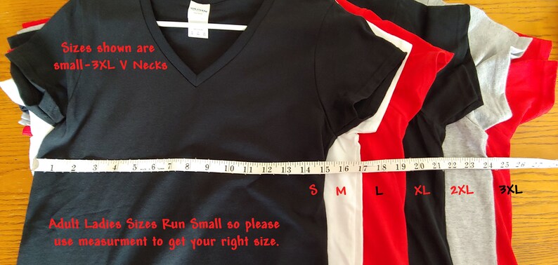 May include: A stack of five V-neck t-shirts in different sizes, from small to 3XL. The shirts are black, white, red, and gray. A measuring tape is laid across the shirts, with the sizes marked in inches. The text "Sizes shown are small-3XL V Necks" and "Adult Ladies Sizes Run Small so please use measurement to get your right size." is printed below the shirts.