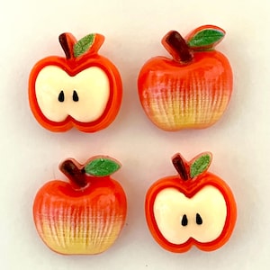 May include: Four apple-shaped decorative embellishments. Two are whole apples, and two are apple halves. The apples are red with yellow gradients, brown stems, and green leaves. The apple halves have white interiors with two black seeds.