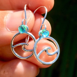 May include: A pair of silver-toned drop earrings featuring a circular frame with an internal wave design. Each earring is accented with a small, translucent blue and green glass bead positioned at the top of the circular wave motif.