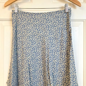 May include: A blue skirt featuring a repeating pattern of small, off-white floral shapes. The skirt has a straight waistband and a slightly flared silhouette, hanging from a wooden hanger with metal clips against a white paneled door.
