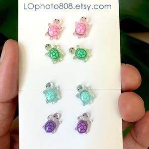 May include: A set of four pairs of turtle-shaped stud earrings in pink, green, blue, and purple. The earrings are displayed on a white card with the text "LOphoto808.etsy.com". The earrings are small and have a glittery appearance.