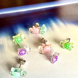 May include: A collection of colorful turtle stud earrings. The earrings are in various shades of pink, green, and purple. Each earring features a small, detailed turtle design with a post backing.