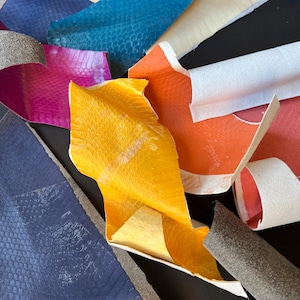 May include: A selection of colourful leather offcuts in shades of blue, teal, pink, orange, and yellow. The leather pieces have a textured, reptile-like pattern and are layered, with some edges curled.