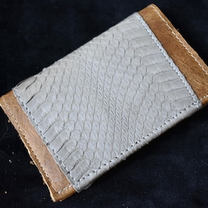 May include: A handmade wallet with a light grey snakeskin-effect central panel, bordered by brown leather edges. The wallet is stitched with white thread, creating a contrast against the darker leather.