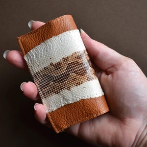 May include: A brown leather card holder with a white and snakeskin patterned centre. The card holder is rectangular and has a stitched border. The snakeskin pattern is a mix of brown and tan.