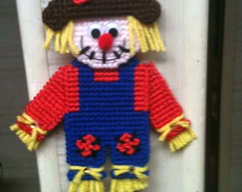 May include: A crocheted scarecrow ornament with a brown hat, red and blue overalls, and yellow straw hair. The scarecrow has a smiling face with a red nose and black eyes.