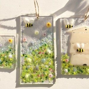 May include: Three glass ornaments featuring a beehive, bees, and flowers. The ornaments are rectangular and have a white background with green, yellow, and blue accents. They are hung with twine.
