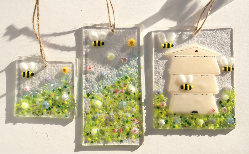May include: Three glass ornaments featuring a beehive, bees, and flowers. The ornaments are rectangular and have a white background with green, yellow, and blue accents. They are hung with twine.
