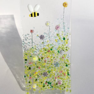 May include: A glass art piece depicting a bee flying over a field of colourful flowers. The flowers are made of small glass pieces in shades of pink, purple, yellow, and green. The bee is made of yellow, black, and white glass.