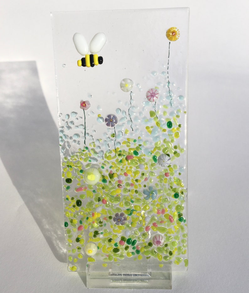 May include: A glass art piece depicting a bee flying over a field of colourful flowers. The flowers are made of small glass pieces in shades of pink, purple, yellow, and green. The bee is made of yellow, black, and white glass.