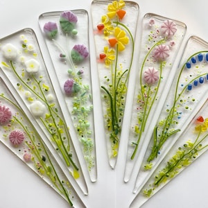 May include: A collection of clear, triangular glass art pieces, each featuring colorful, fused glass floral designs. The flowers include pink, white, yellow, and blue blooms with green stems and leaves. The pieces are arranged in a fan shape.