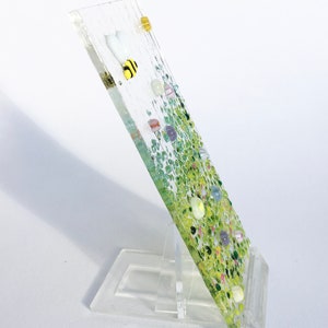 May include: A glass art piece with a green and white background and a bee design. The piece is on a clear stand.