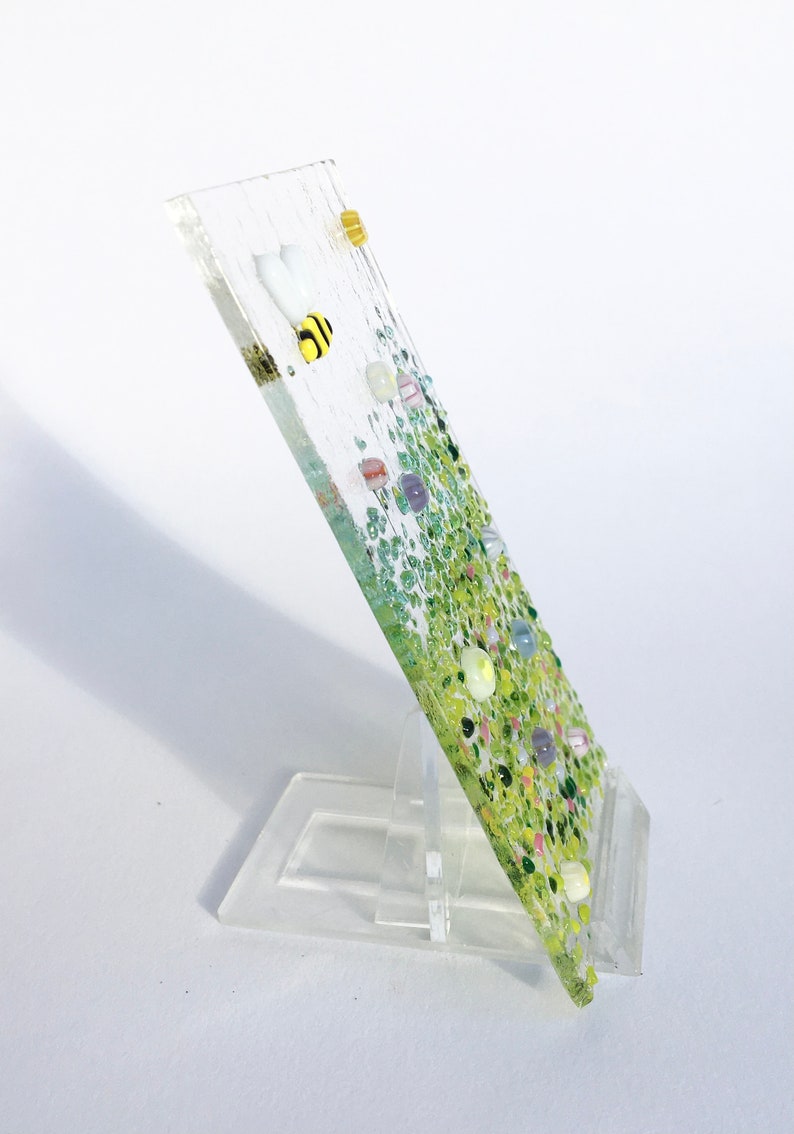 May include: A glass art piece with a green and white background and a bee design. The piece is on a clear stand.