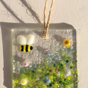 May include: A glass suncatcher featuring a yellow and black bee, green grass, and colourful flowers. The suncatcher is suspended from a twine string.