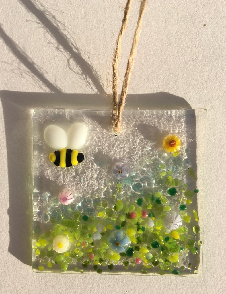 May include: A glass suncatcher featuring a yellow and black bee, green grass, and colourful flowers. The suncatcher is suspended from a twine string.