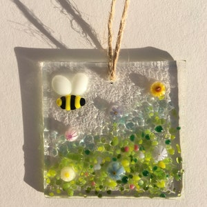 May include: A glass suncatcher featuring a bee and flowers. The bee is yellow and black with white wings. The flowers are yellow, pink, blue, and white. The suncatcher is suspended from a string.