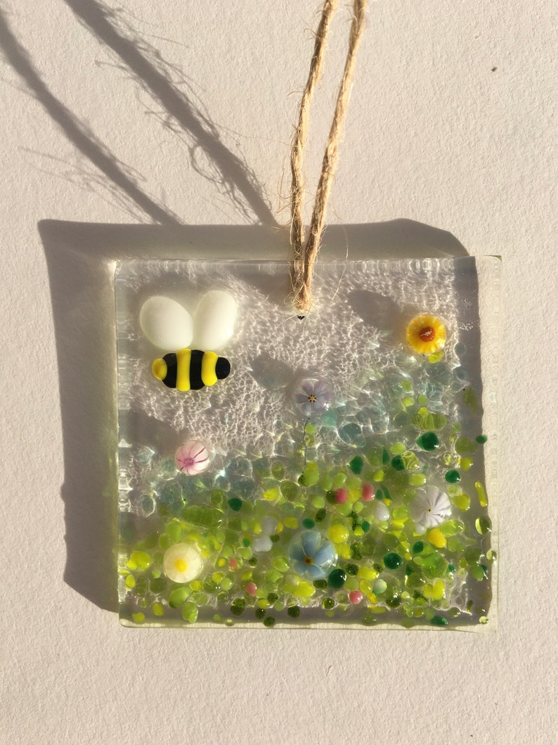 May include: A glass suncatcher featuring a bee and flowers. The bee is yellow and black with white wings. The flowers are yellow, pink, blue, and white. The suncatcher is suspended from a string.