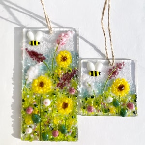 May include: Two glass art pieces with floral designs and bees. One is rectangular, the other square. Both feature yellow sunflowers, pink and purple flowers, and green foliage. Each has a small bee and a twine loop for hanging.